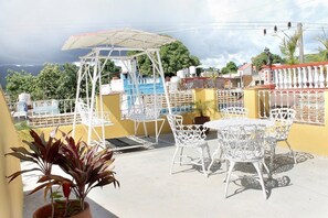 An outdoor patio with white metal chairs and tables, a covered seating area, and a view of the surrounding landscape. - Hostal Juan Fernandez (Trinidad)