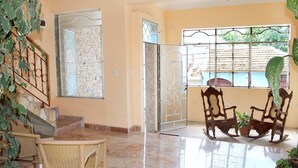 A spacious room with a tiled floor, a dining area with wicker chairs, a rocking chair, and a potted plant. - Hostal Juan Fernandez (Trinidad)