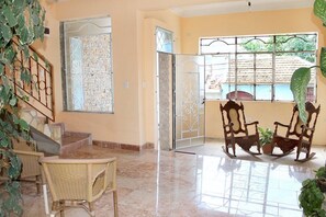 A spacious room with a tiled floor, a dining area with wicker chairs, a rocking chair, and a potted plant. - Hostal Juan Fernandez (Trinidad)