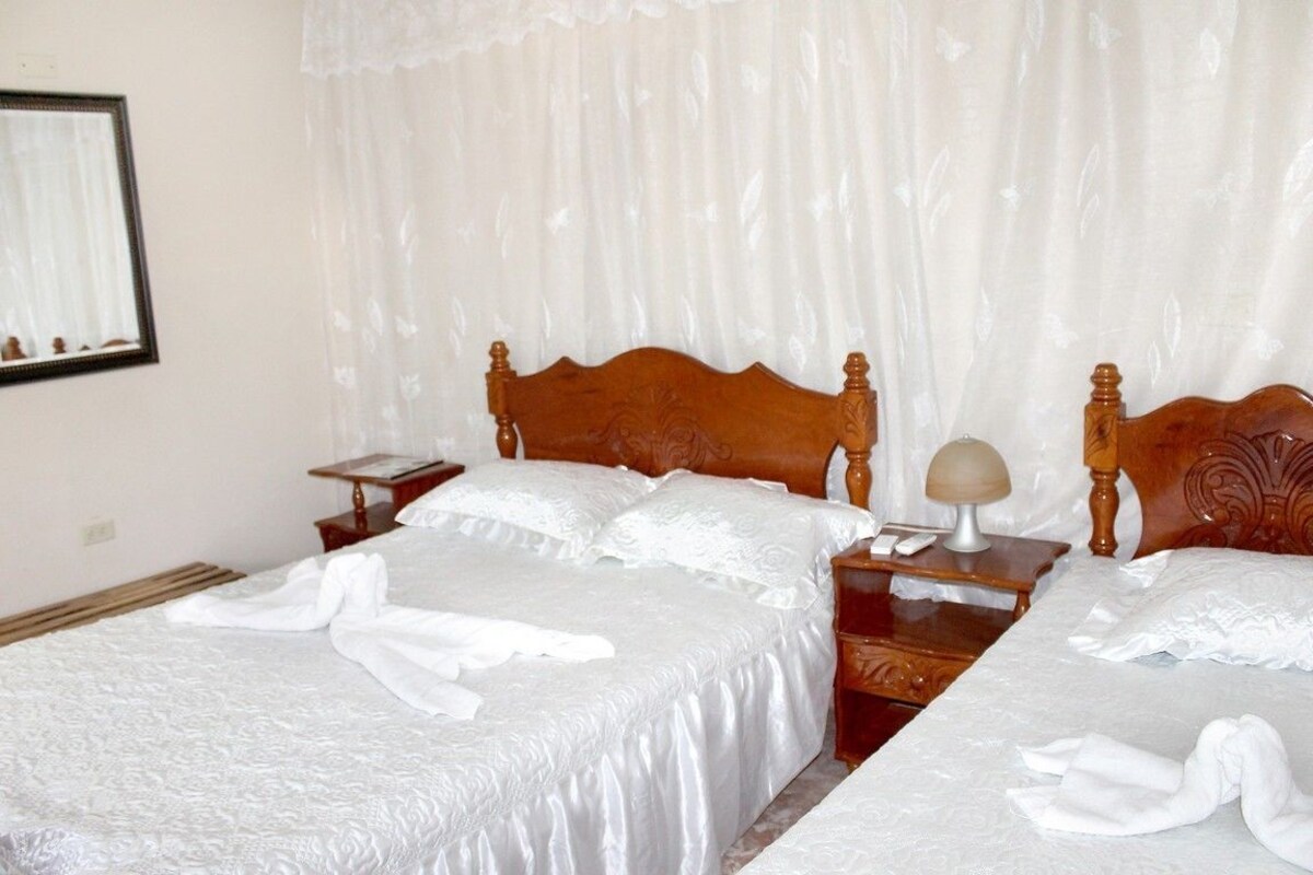 A hotel room with two beds, each with a wooden headboard and white linens. There is a nightstand with a lamp between the beds.