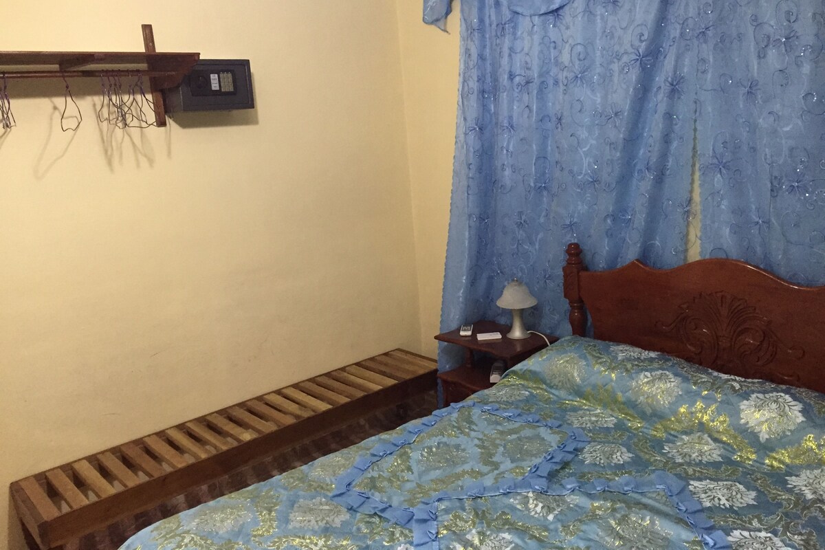 A bedroom with a wooden bed, a nightstand with a lamp, a small shelf with items, and a blue curtain.