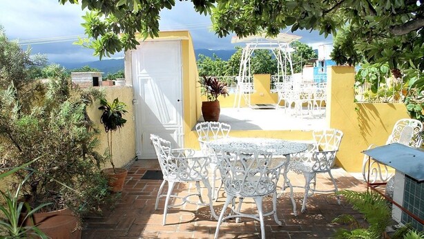 An outdoor patio with white metal chairs and a table, surrounded by potted plants and a yellow wall. - Hostal Juan Fernandez (Trinidad)