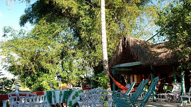 Mobiliário ao ar livre com mesas e cadeiras, estrutura com telhado de palha e vegetação exuberante ao fundo.