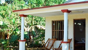 Uma área de estar coberta ao ar livre, com duas cadeiras de madeira, um edifício branco com telhado vermelho e uma janela com grades.