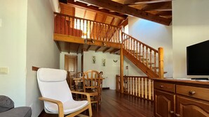 Un vaste espace de vie comprenant un escalier en bois, une table à manger avec des chaises, un fauteuil blanc et un téléviseur posé sur un meuble en bois.