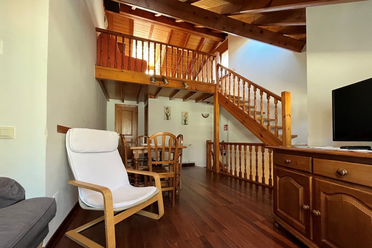 Een ruime woonkamer met een houten trap, een eettafel met stoelen, een witte fauteuil en een tv op een houten kast.