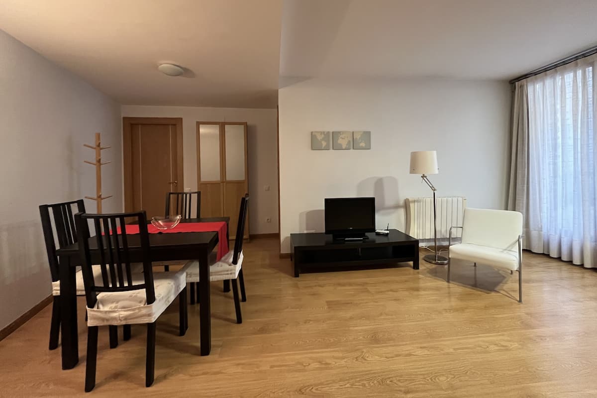 Een woonkamer met een eettafel, stoelen, een televisie, een lamp en een groot raam met gordijnen.