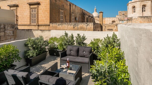 A rooftop terrace with outdoor furniture, potted plants, and a view of historic buildings. - Valletta Kampnar City Living (Valletta)