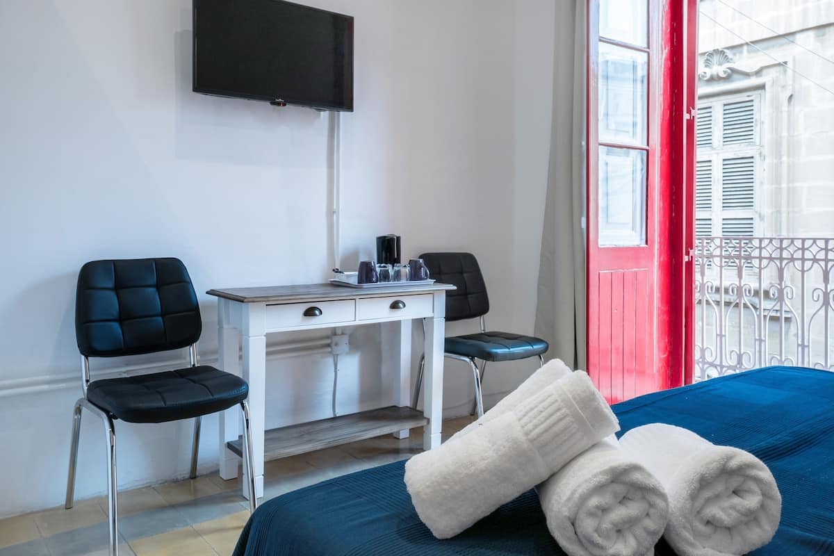 A hotel room with a bed, two chairs, a desk, a flat-screen TV, and a window with a red door.