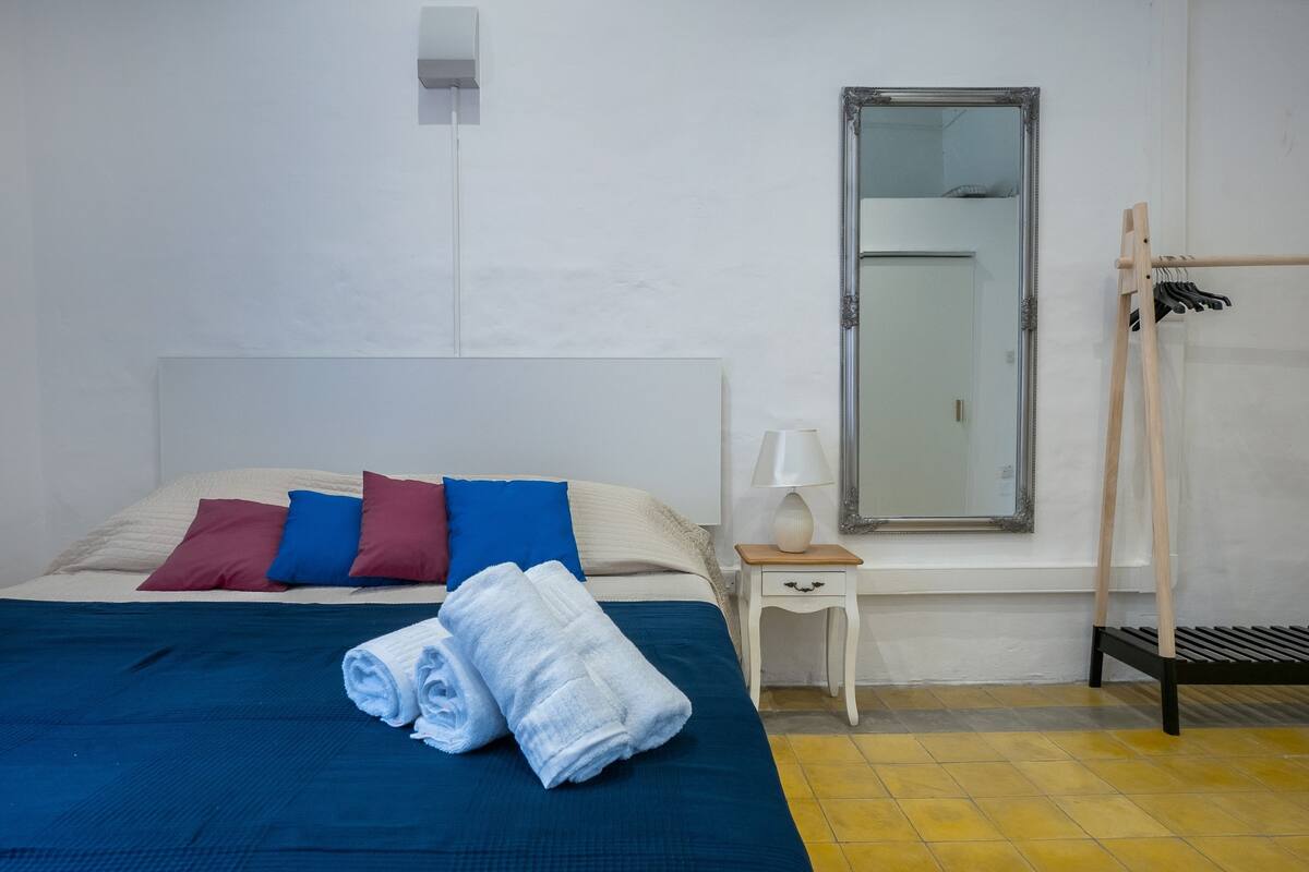 A neatly made bed with a blue bedspread and rolled white towels, a bedside table with a lamp, a mirror, and a wooden ladder.