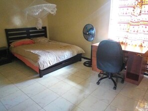Une chambre à coucher comprenant un lit, un bureau, une chaise, un ventilateur sur pied et une fenêtre avec des rideaux.