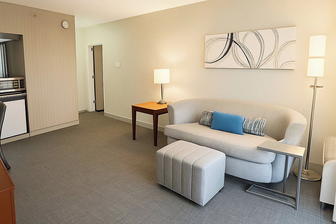 Ein modernes Hotelzimmer mit grauem Sofa, Hocker, kleinem Tisch und Lampe. Im Hintergrund ist eine Küchenzeile erkennbar und eine Tür zu einem weiteren Raum.