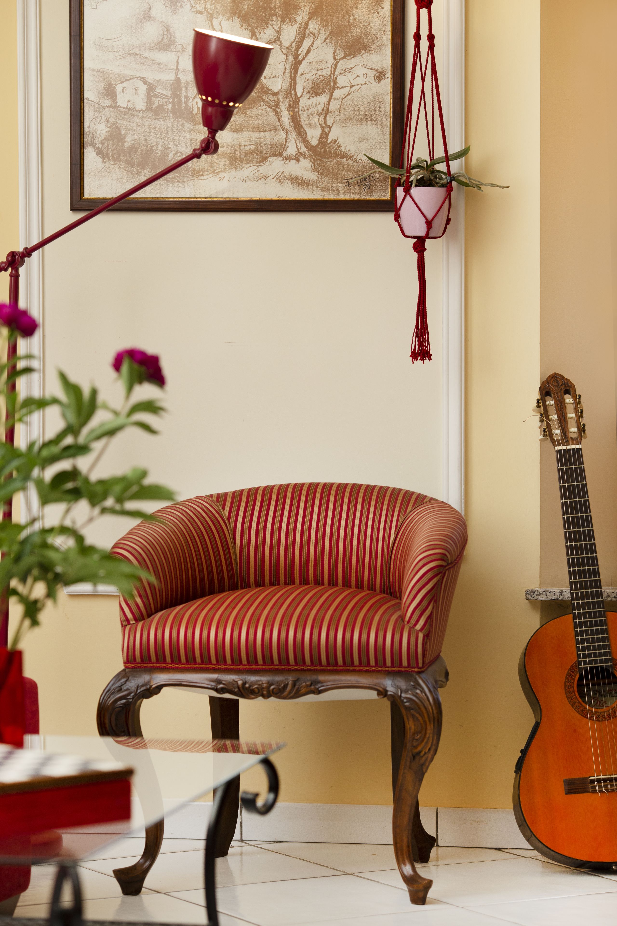 Ein Zimmer mit einem rot gestreiften Sessel, einer Gitarre, einer hängenden Zimmerpflanze aus Makramee und einem gerahmten Landschaftsbild an der Wand.