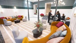 A modern hotel lobby with a curved yellow sofa, a grey armchair, and a striped ottoman. There is a black coffee table with a vase of flowers. The floor has a striped pattern, and the walls are painted in teal and white. - Soho Boutique Congreso (Madrid)