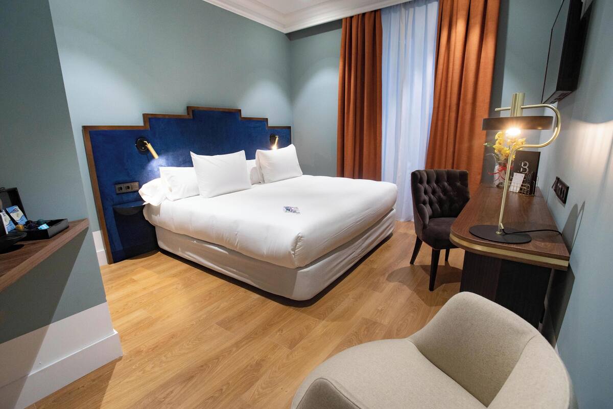 A hotel room with a large bed, a desk with a lamp, a chair, and a wooden floor.