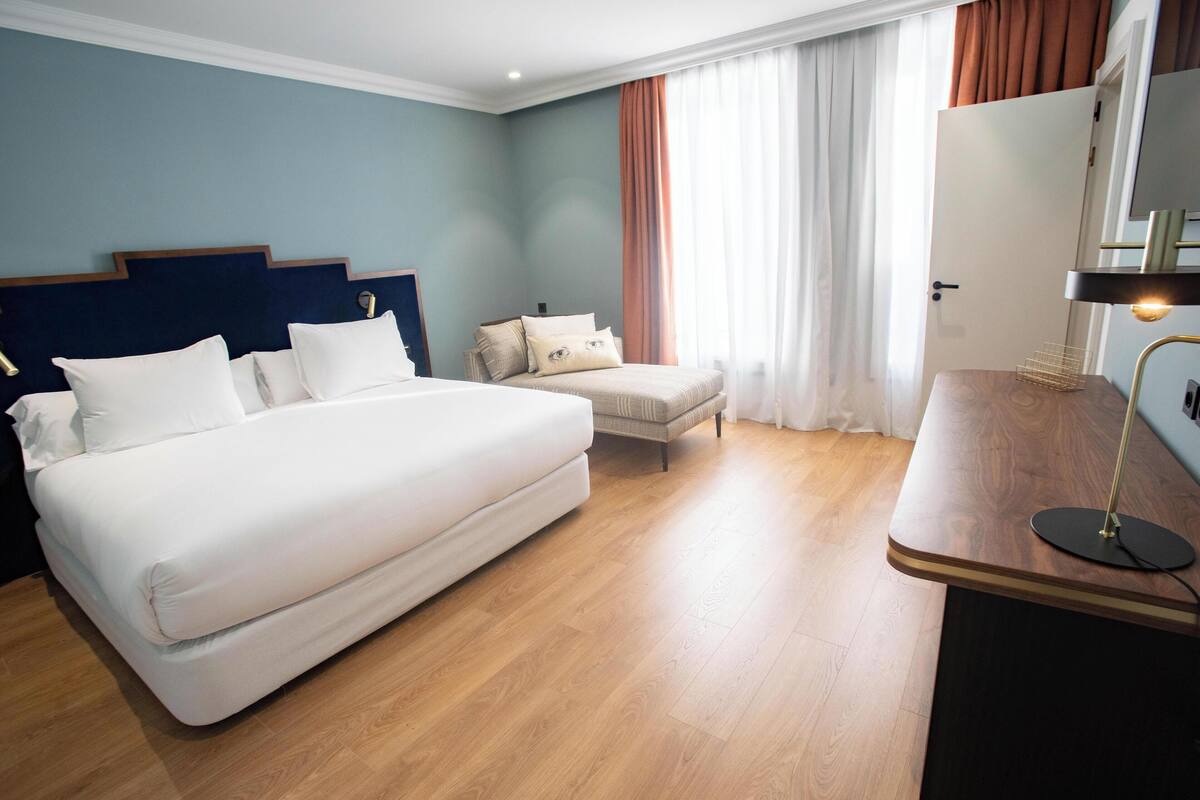 A hotel room with a large bed, a bedside table with a lamp, a small chair, and a desk with a mirror.