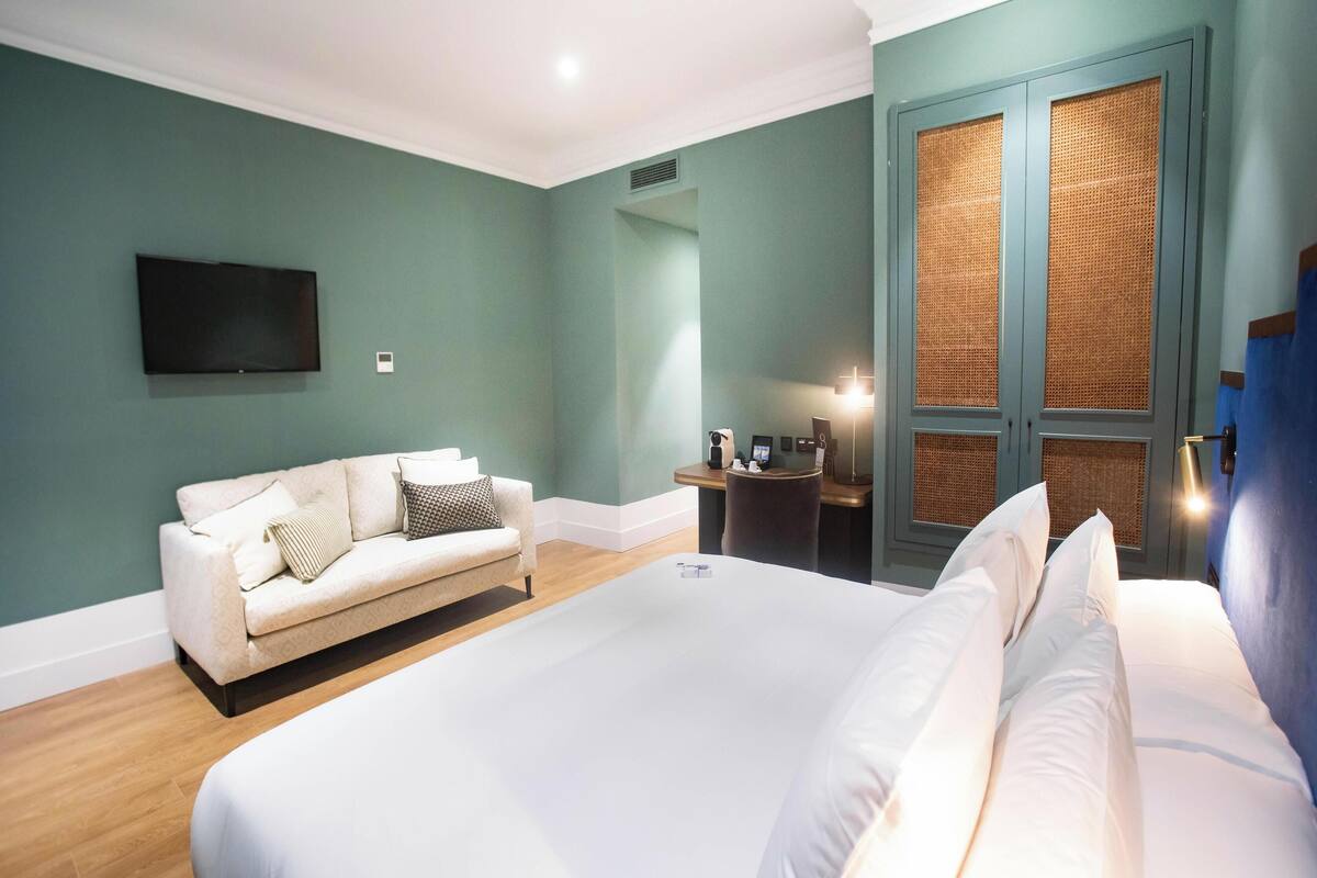 A hotel room with a large bed, a sofa, a TV mounted on the wall, a small table with a lamp, and a window with a view.