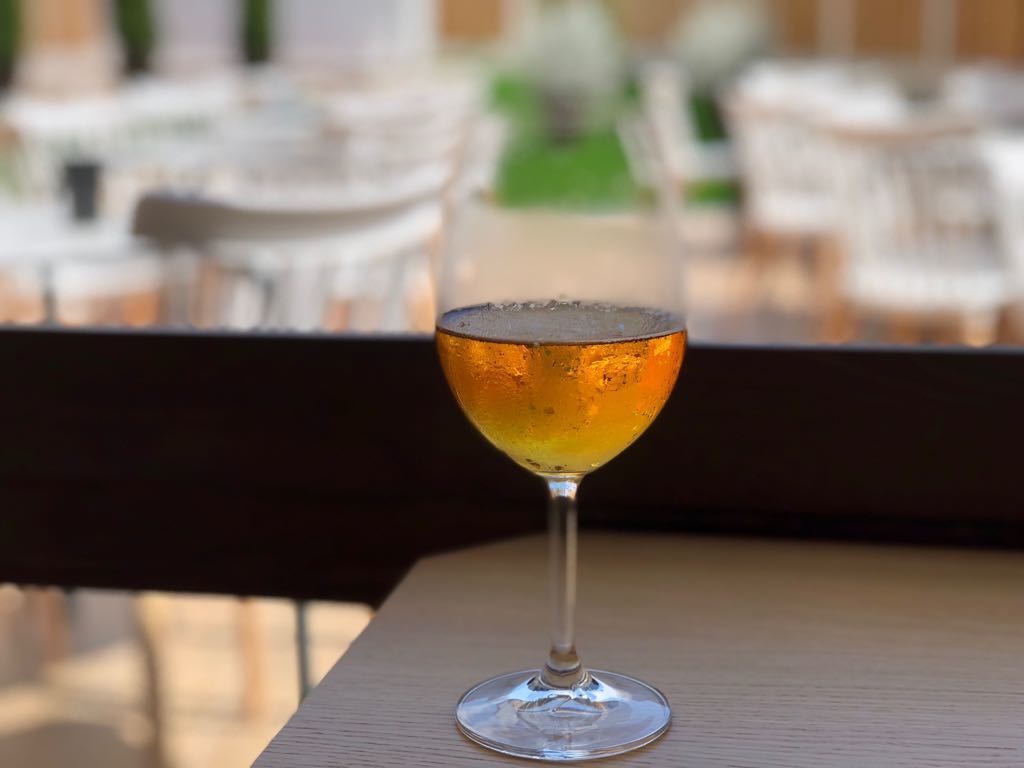 Un vaso de cóctel sobre una mesa de madera, con un área de asientos al aire libre desenfocada al fondo.