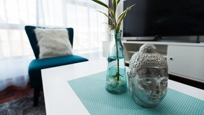 Uma sala de estar moderna com um sofá azul-turquesa, uma mesa de centro branca, um vaso de vidro com uma planta e um busto decorativo.