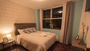 Uma cama bem arrumada com cabeceira, mesa de cabeceira com abajur e janela com cortinas.