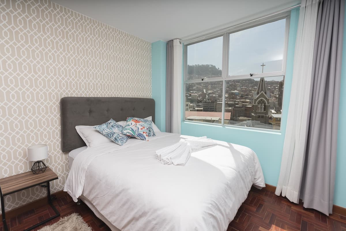 A bedroom with a large bed, a nightstand, a lamp, and a view of a cityscape through a window.