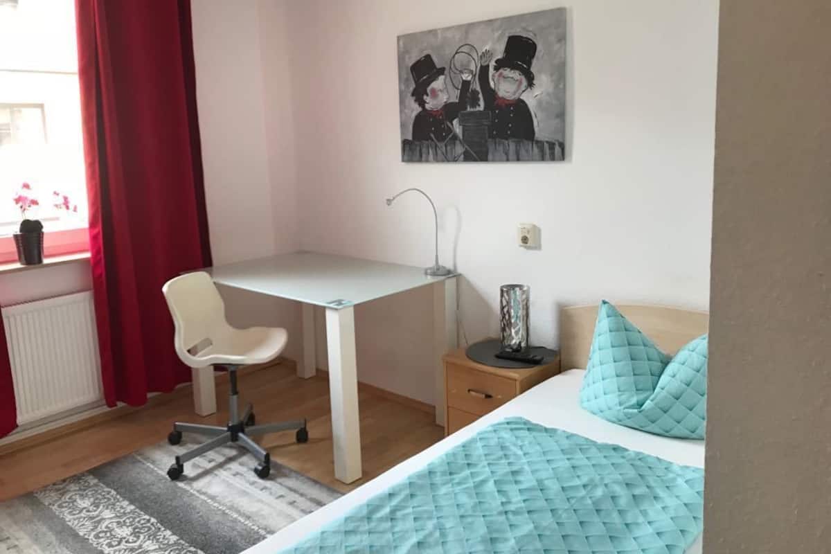 A bedroom with a bed, a desk, a chair, a nightstand, and a window with red curtains.