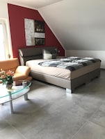 Quarto moderno com cama, mesa de centro de vidro, sofá e parede de destaque vermelha.