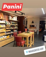 Uma loja Panini com prateleiras cheias de lanches e bebidas, uma mesa de madeira com cadeiras vermelhas e uma lareira ao fundo.