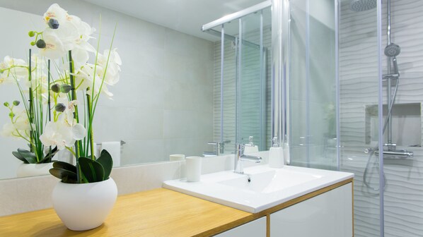 A modern bathroom with a white sink, mirror, and shower. There is a potted orchid on the counter.