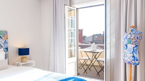 Ein Hotelzimmer mit Bett, Nachttisch mit Lampe, Balkon mit Tisch und Stühlen und Blick auf Gebäude.