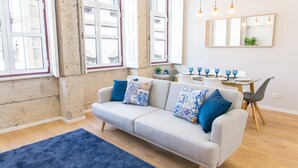 A modern living room with a light grey sofa, blue patterned cushions, a blue rug, and a dining area with blue glassware. - Praça 44 Boutique Apartments (Braga)
