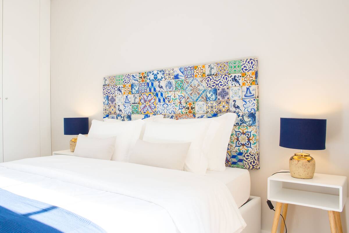 A neatly made bed with white linens, a colorful tile headboard, and two bedside lamps on white nightstands.