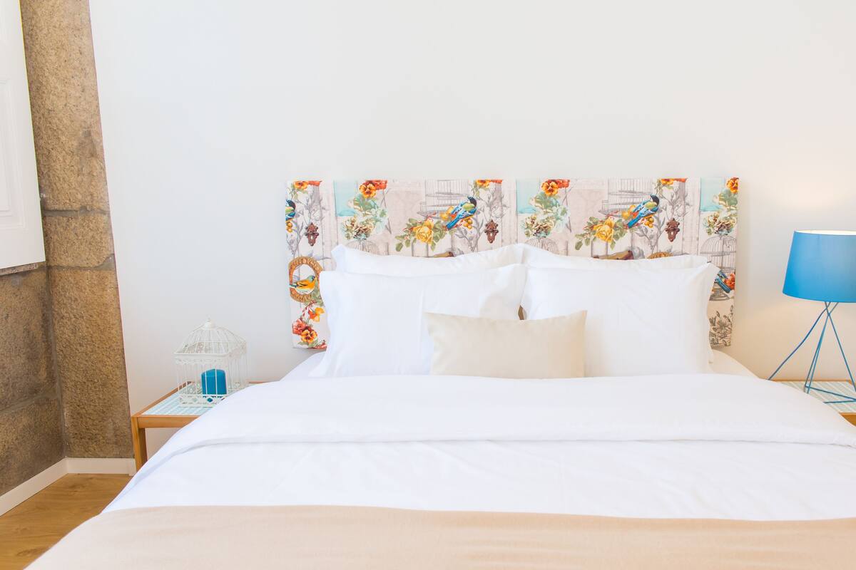 A neatly made bed with white linens, a floral headboard, a small bedside table with a lamp, and a wall with a bird-themed wallpaper.