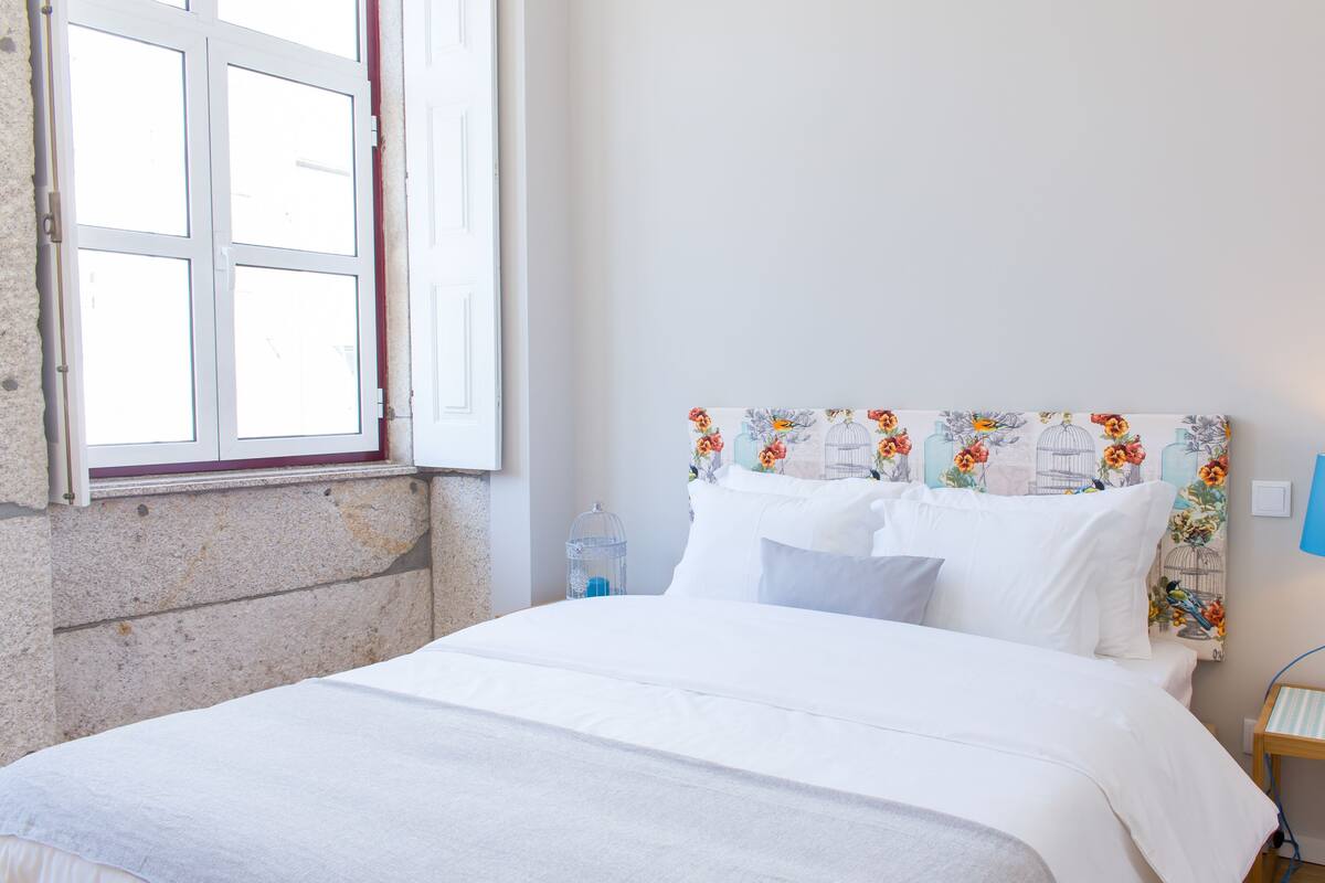 A neatly made bed with white linens, a headboard with floral patterns, a bedside table with a lamp, and a window with a view of the outside.