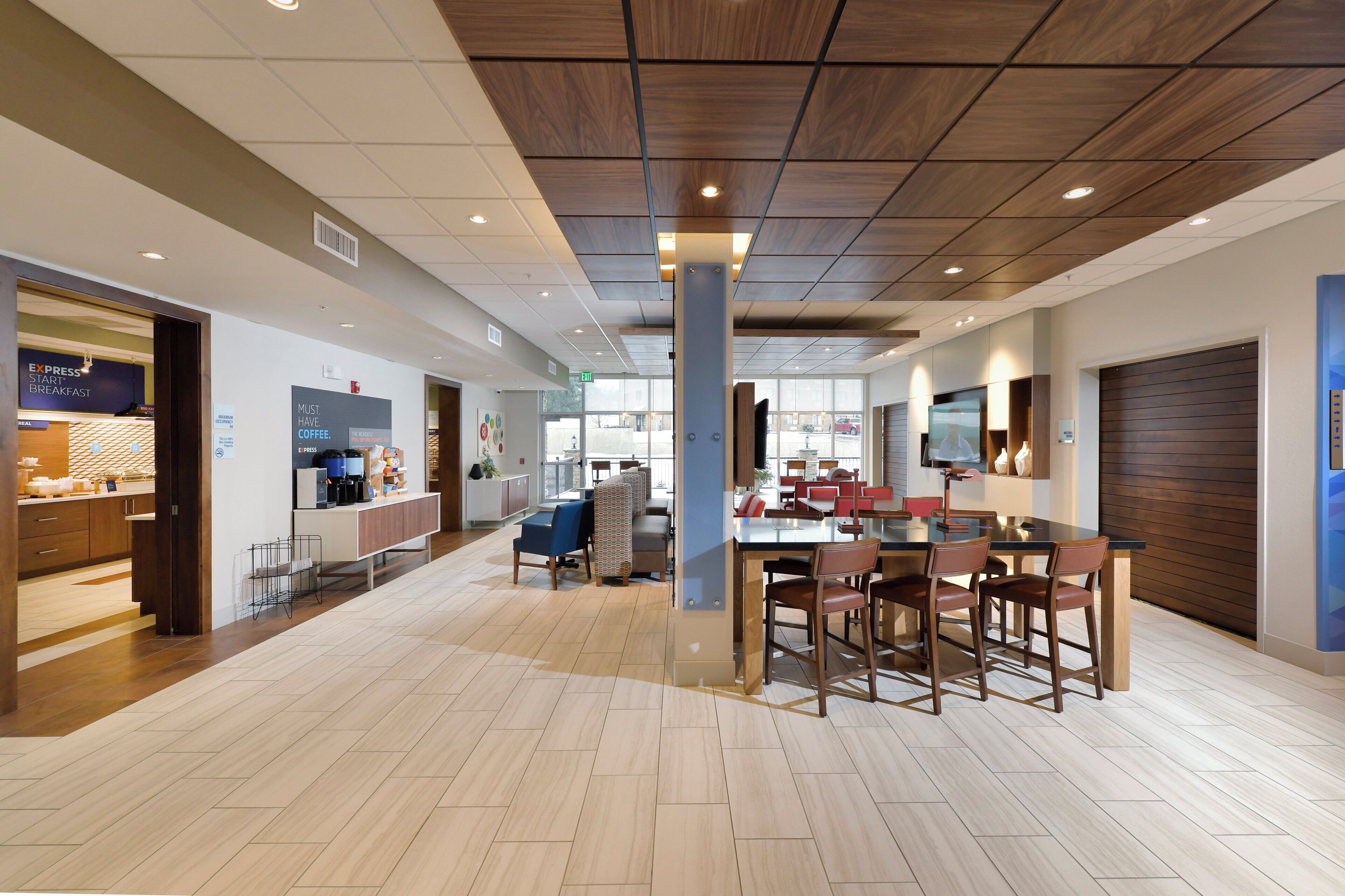 A modern cafe interior with a wooden ceiling, a central dining table with chairs, and a visible espresso machine.