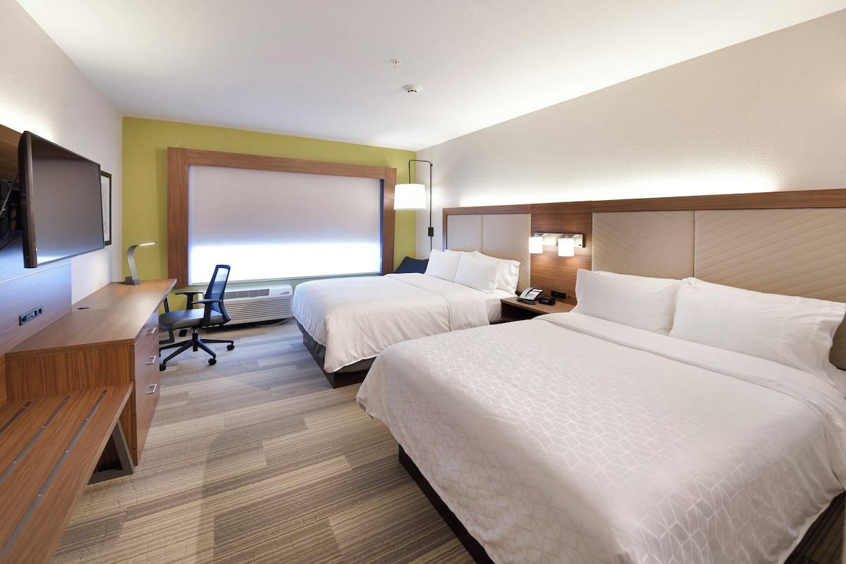 Une chambre d’hôtel avec deux lits, un bureau avec une chaise, un téléviseur à écran plat et une fenêtre avec des stores.