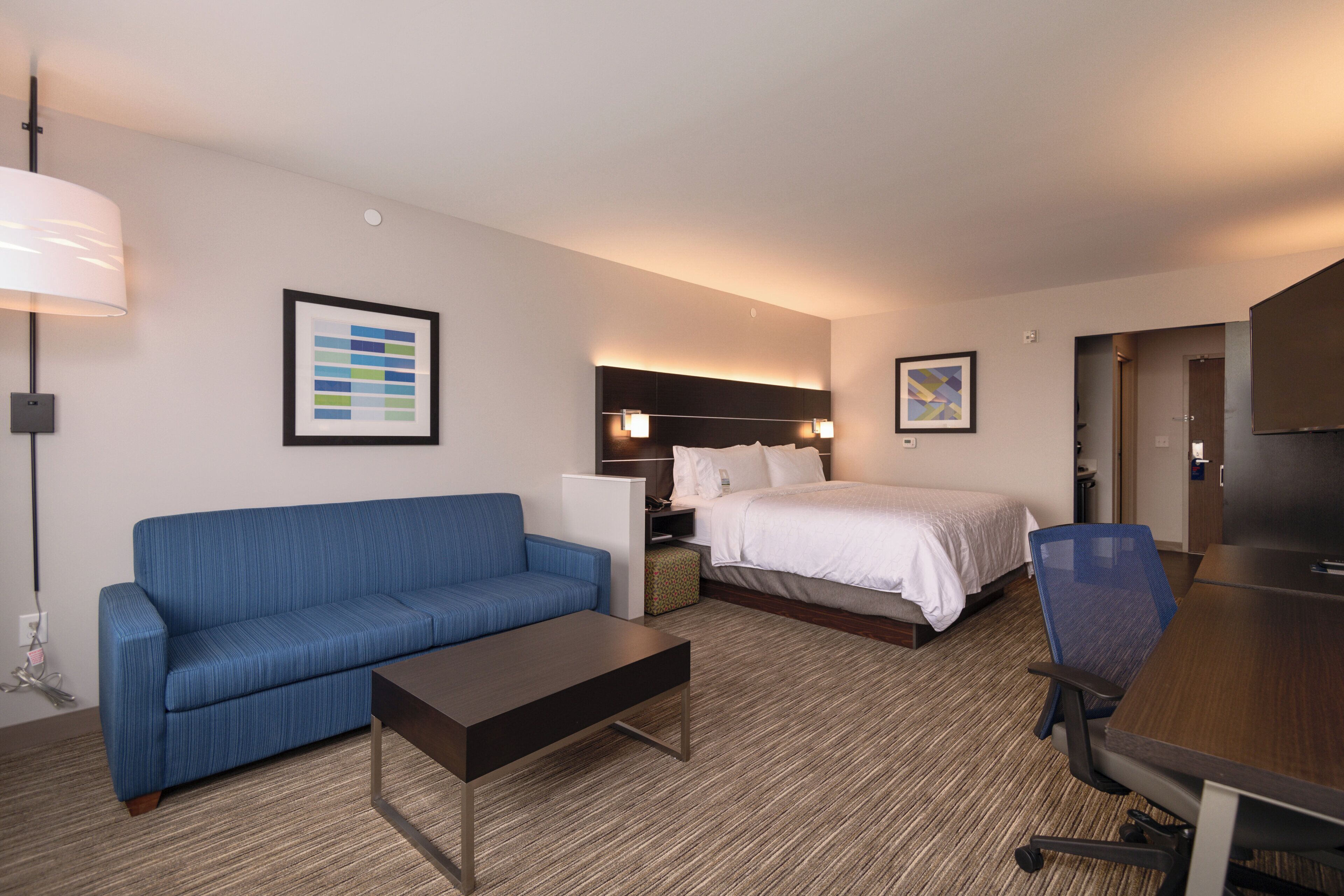 Quarto de hotel com sofá azul, cama com roupa de cama branca, escrivaninha e cadeira, e televisão.
