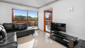 A spacious living room with a black leather sofa, a glass coffee table, and a flat-screen TV. Large windows offer a view of the outdoors. - San Lameer Villa Rentals 10413 (Southbroom)