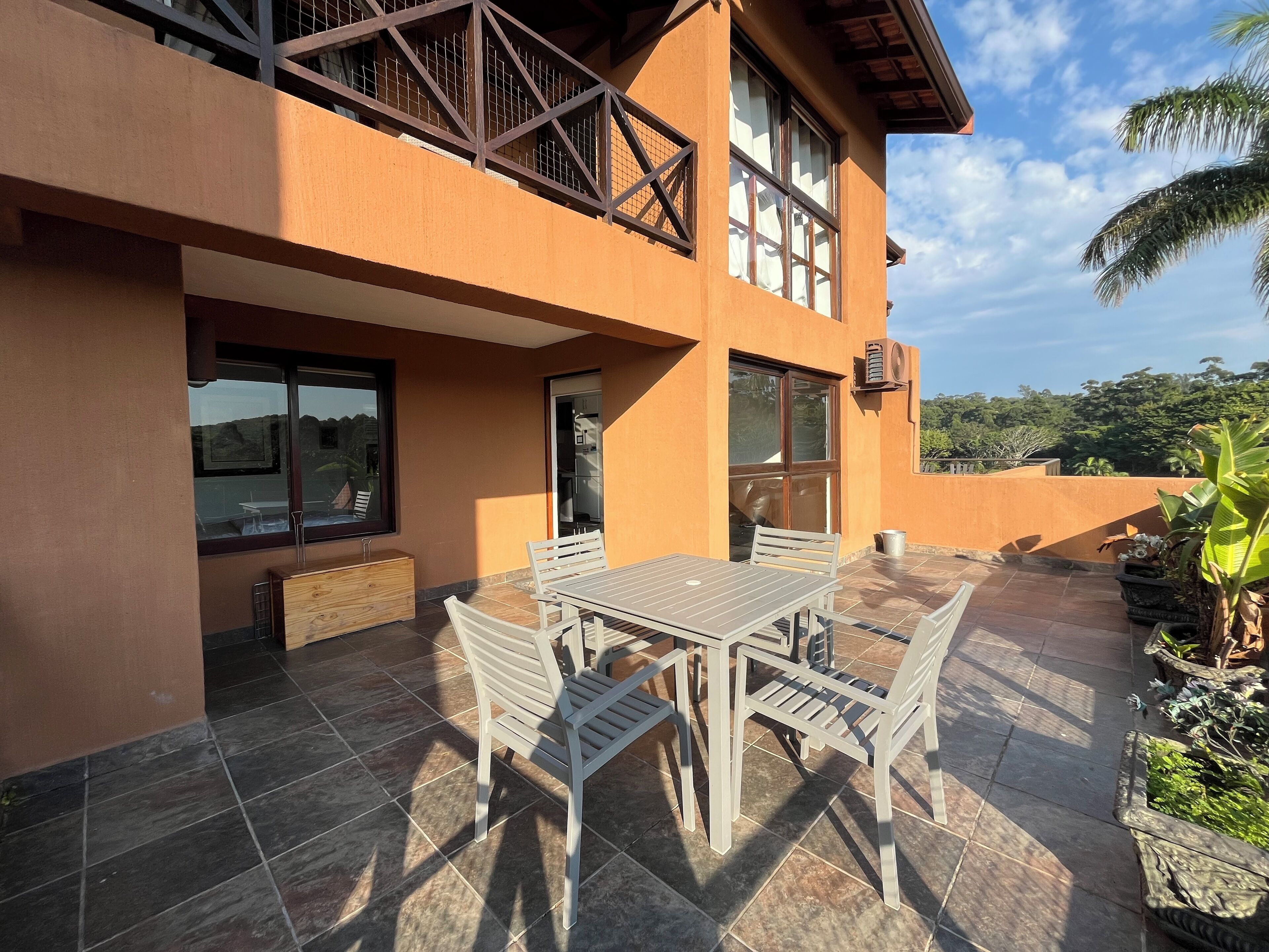 A modern two-story building with a patio area featuring a table and chairs, a wooden bench, and potted plants.