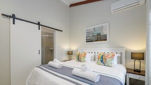 A neatly made bed with a striped bedspread, two pillows with a floral pattern, and two rolled towels. There is a bedside table with a lamp, a wall-mounted air conditioner, and a framed picture above the bed. - San Lameer Villa Rentals 2204 (Southbroom)