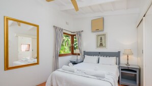 A neatly made bed with white linens, a bedside table with a lamp, a nightstand, a large mirror, and a window with curtains. - San Lameer Villa Rentals 2408 (Southbroom)