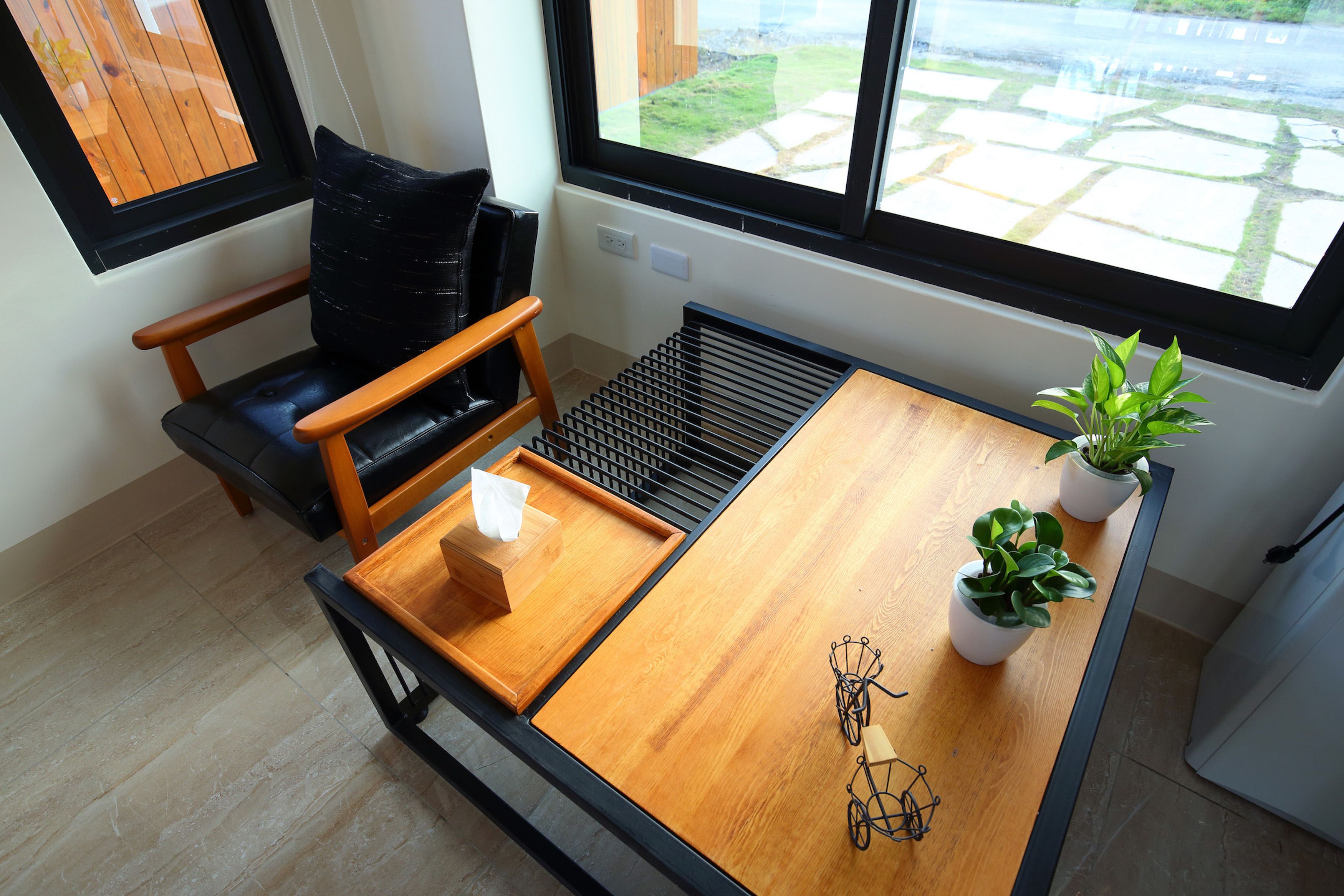 A compact room with a wooden table, a black chair, a small potted plant, and a window with a view of a garden.