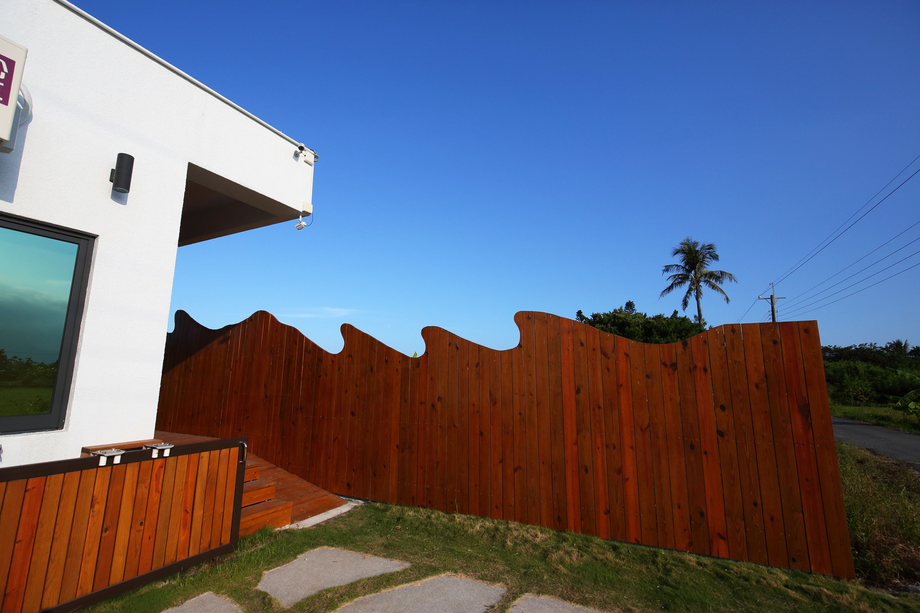 A modern house with a wooden fence, a paved pathway, and a palm tree in the background.