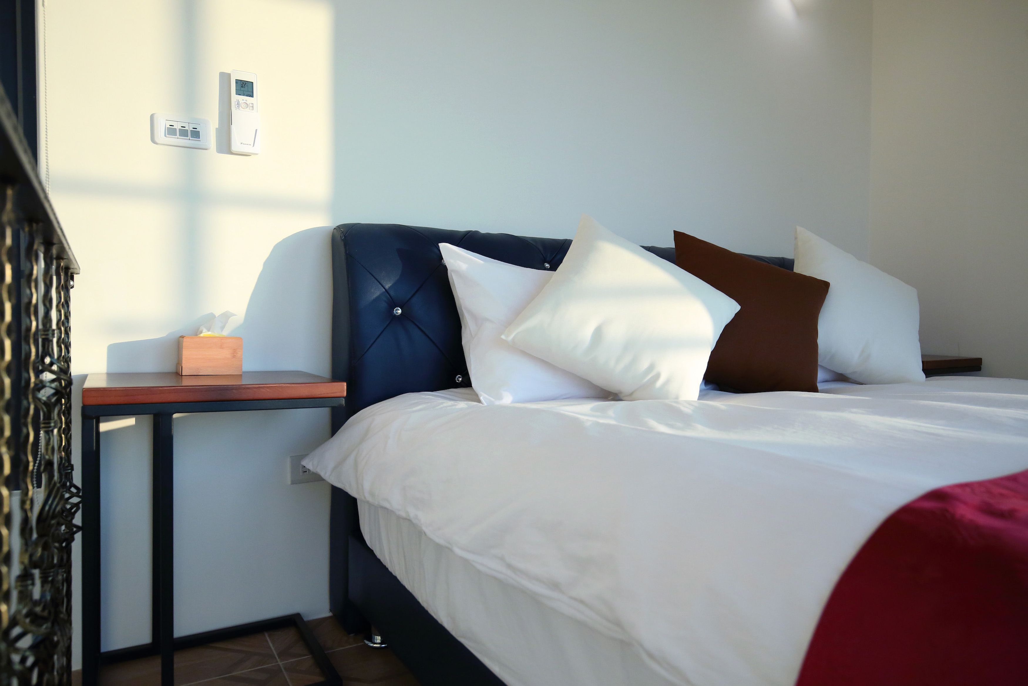 A neatly made bed with white bedding and multiple pillows, a bedside table with a tissue box, and a wall-mounted thermostat.