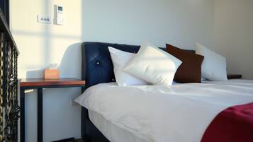 A neatly made bed with white bedding and multiple pillows, a bedside table with a tissue box, and a wall-mounted thermostat.
