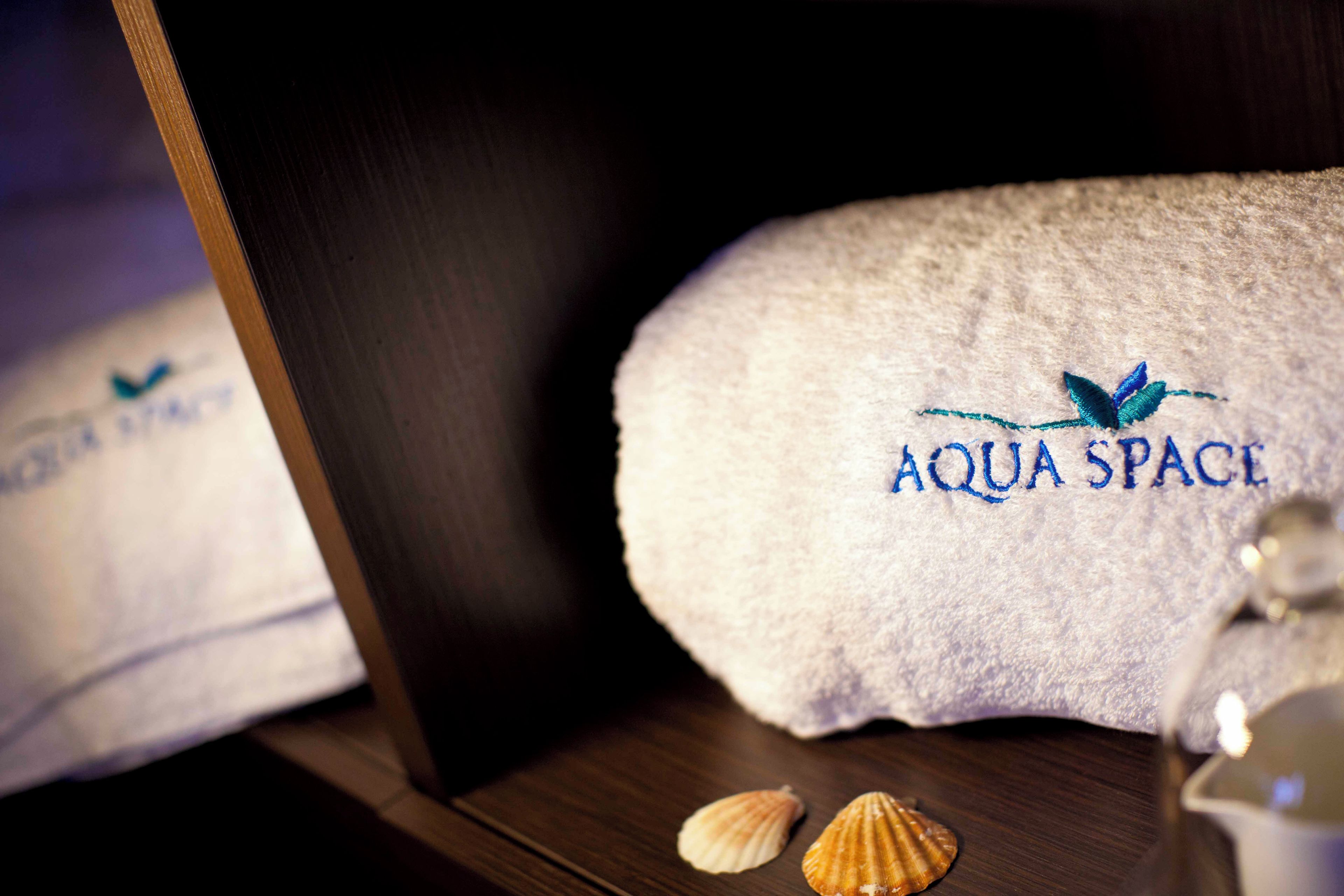 A towel with 'AQUA SPACE' embroidered on it, placed on a wooden surface with two seashells nearby.