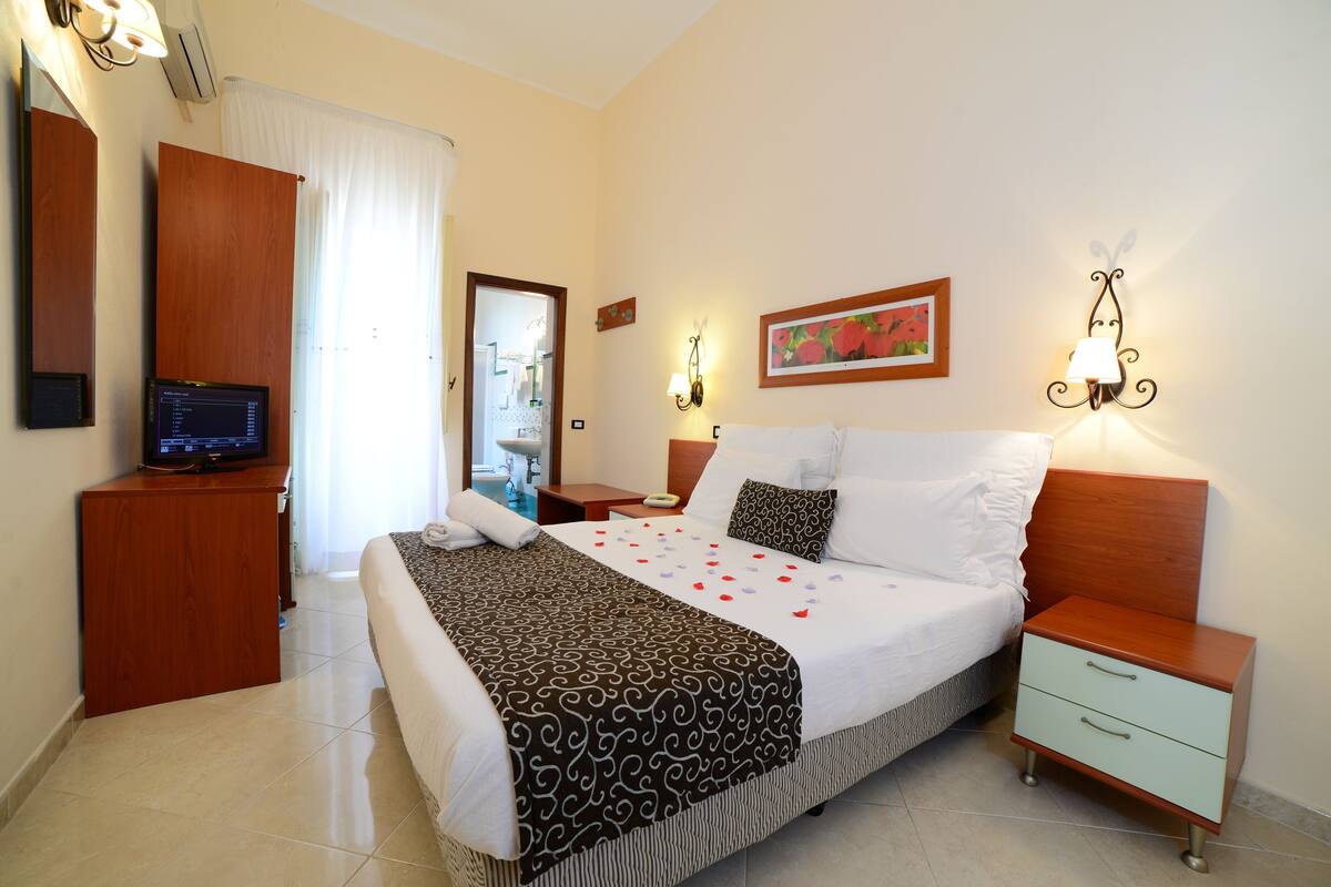 A neatly arranged hotel room with a bed, bedside tables, a television, and a bathroom visible through an open door.