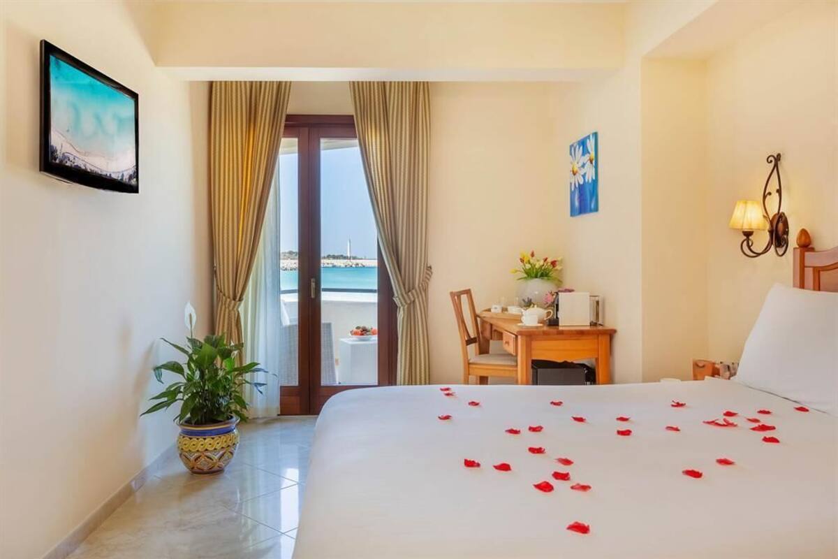 A hotel room with a bed, a desk, a chair, a potted plant, and a view of the sea through a door.