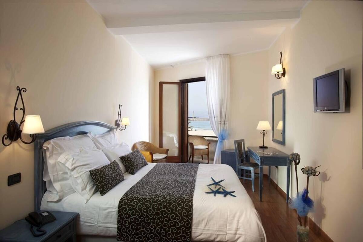A hotel room with a bed, bedside lamps, a desk, a chair, a TV, and a view of the sea.