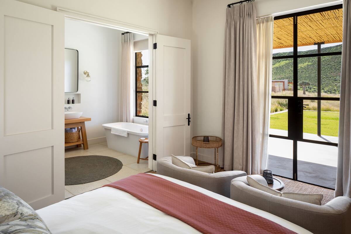 A bedroom with a large bed, a sitting area, a bathroom, and a view of the outdoors.